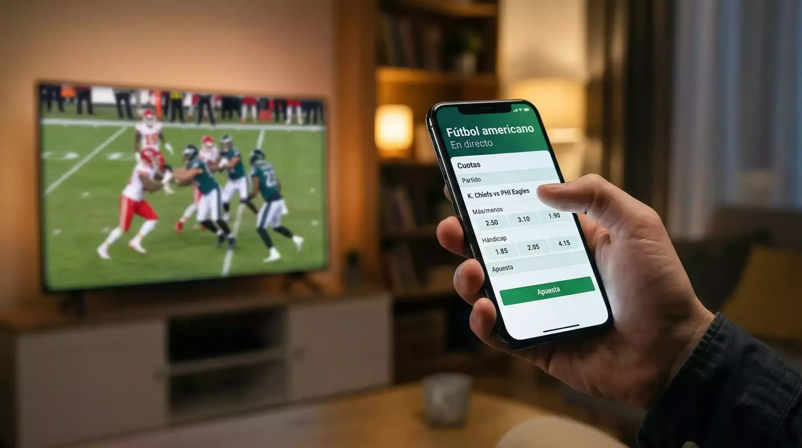 Mano sosteniendo un teléfono móvil con la pantalla encendida frente a un televisor con fútbol americano